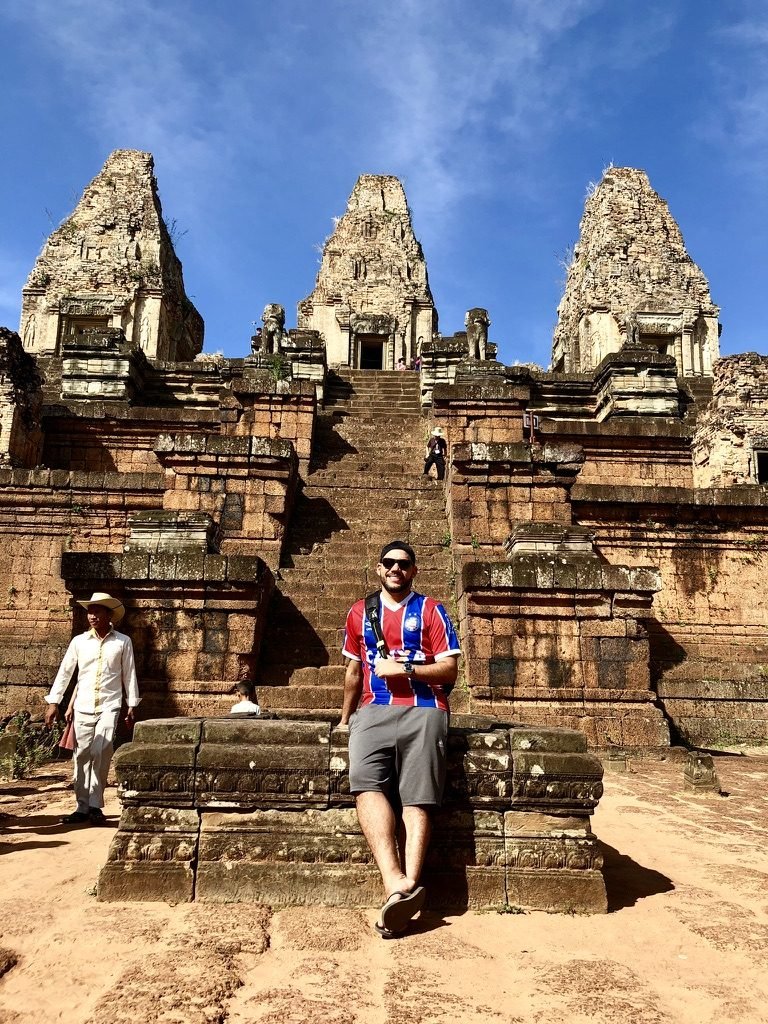 Tiago Lopes sentado na base de um templo de tijolos com três torres (Pre Rup), vestindo a camisa do Bahia