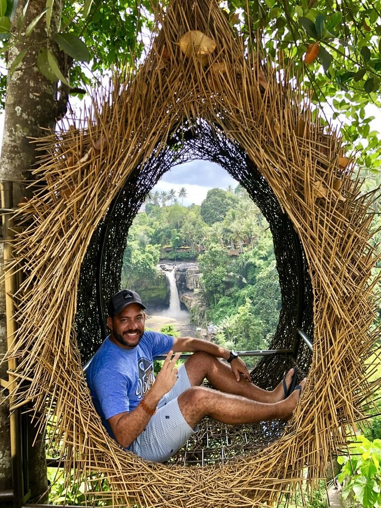 Tiago Lopes sentado em um ninho de palha com uma cachoeira ao fundo, em meio à floresta