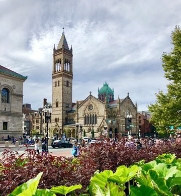 Igreja de pedra com torre alta em uma praça de Boston, com canteiros floridos em primeiro plano