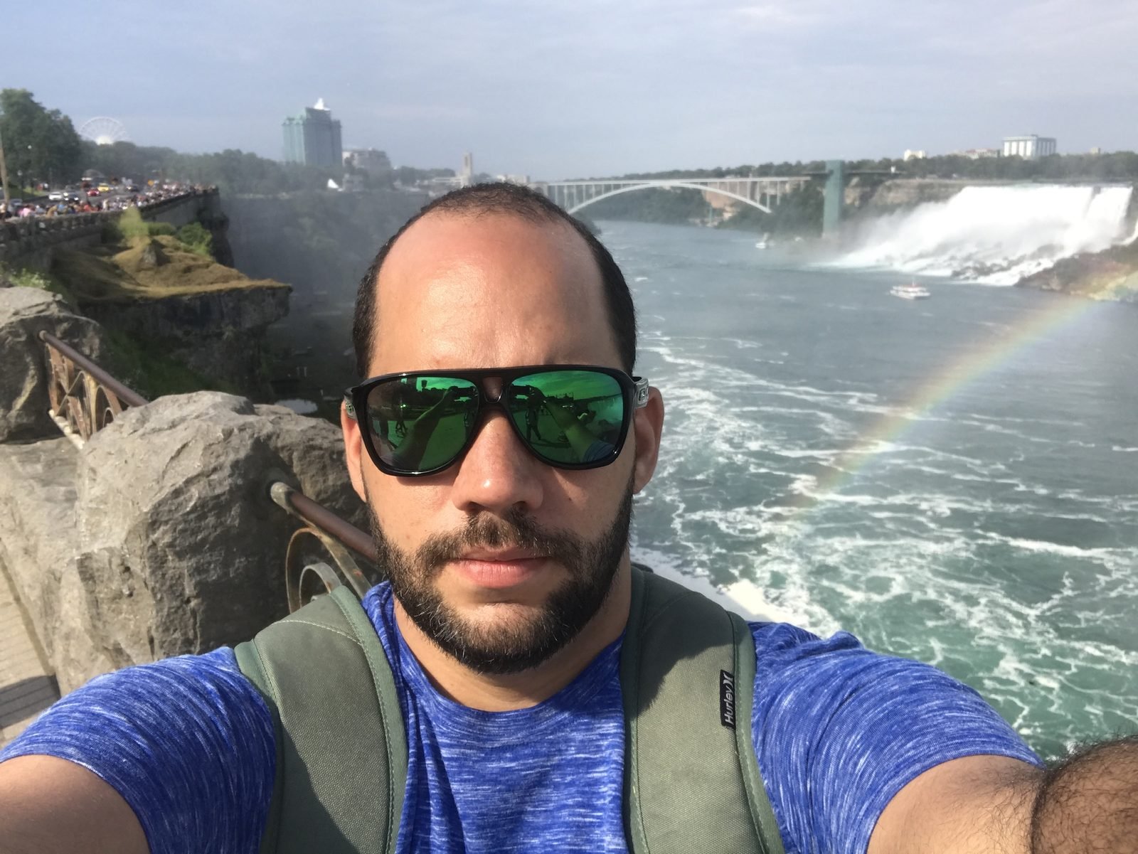 Tiago Lopes de óculos escuros diante das Cataratas do Niágara, com um arco-íris sobre a água