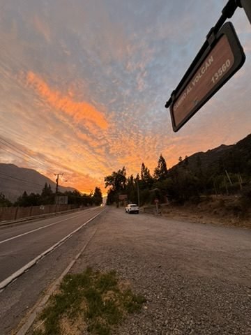 Céu alaranjado ao entardecer sobre a estrada, com a placa Camino al Volcán