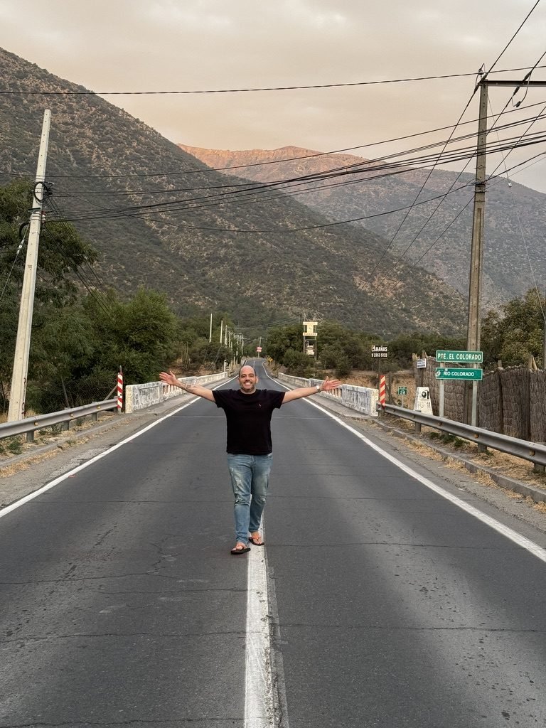 Tiago Lopes de braços abertos no meio de uma estrada de montanha no Cajón del Maipo, com a placa Puente El Colorado ao fundo