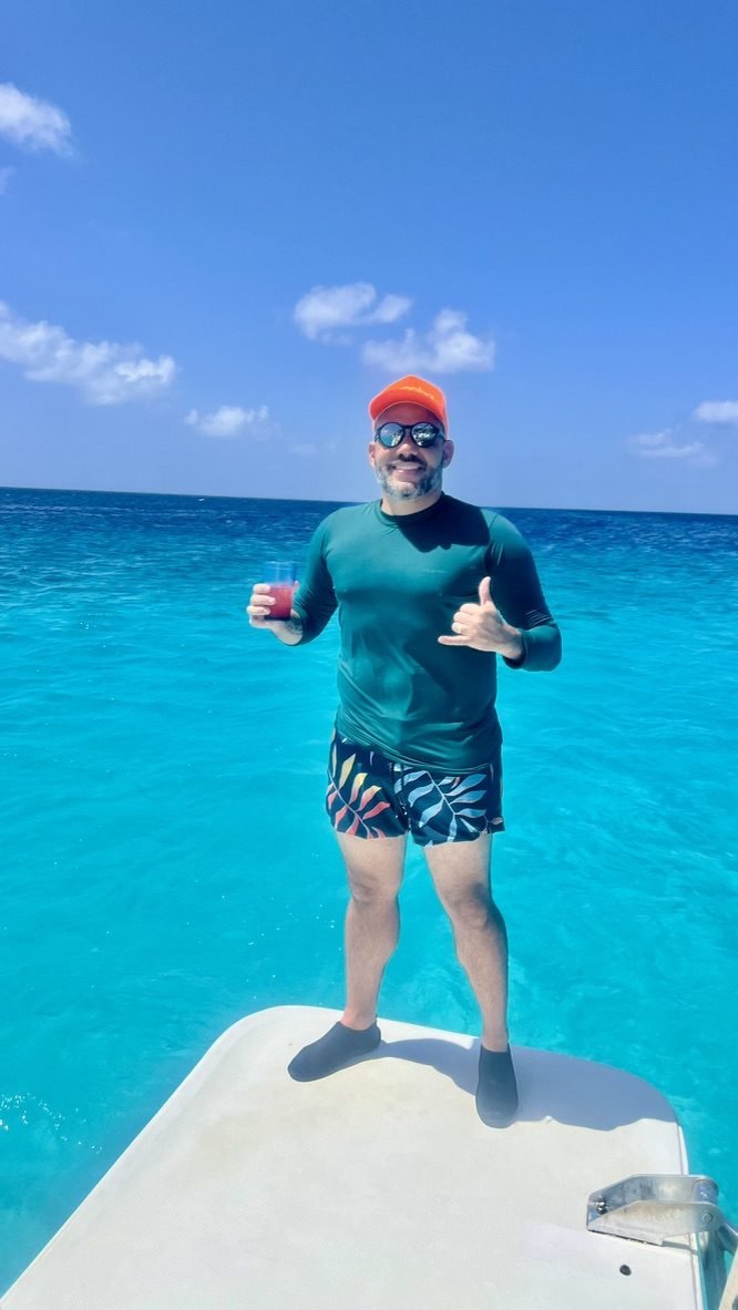 Tiago Lopes na proa de um barco com um drink na mão, sobre o mar azul-turquesa de Curaçao