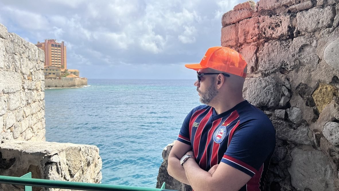 Tiago Lopes de camisa do Bahia encostado na muralha de um forte, olhando o mar