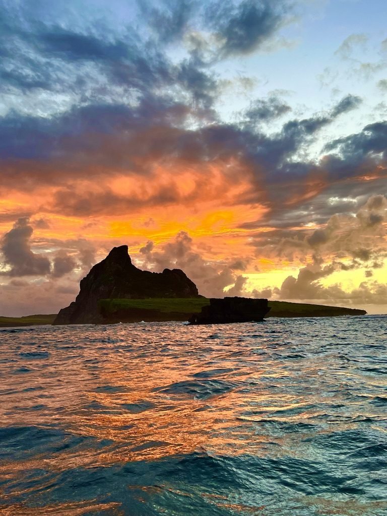 Pôr do sol alaranjado sobre o mar, com uma ilha rochosa em silhueta
