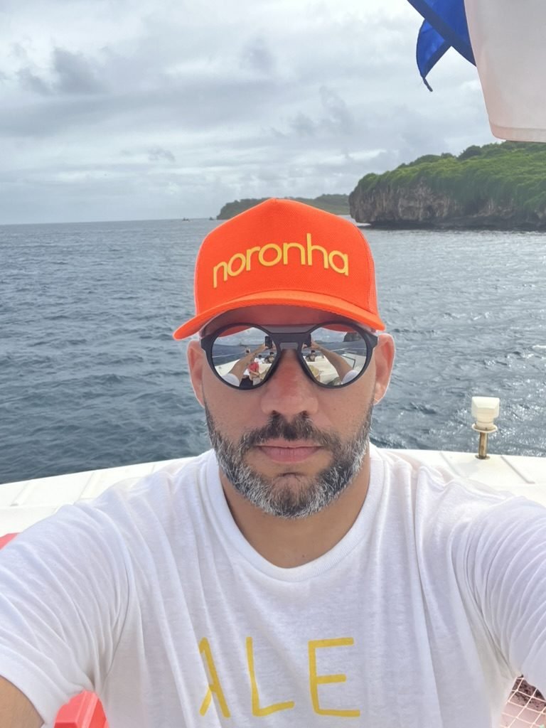 Tiago Lopes em selfie no barco com um boné laranja escrito Noronha