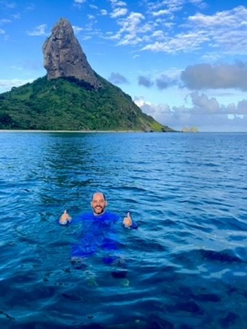 Tiago Lopes boiando no mar azul com os polegares para cima, com o Morro do Pico ao fundo