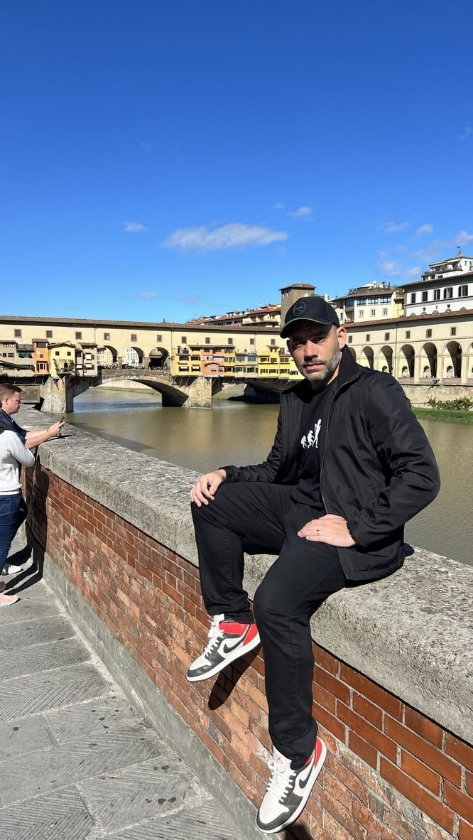 Tiago Lopes sentado na mureta à beira do rio Arno, em Florença, com a Ponte Vecchio ao fundo
