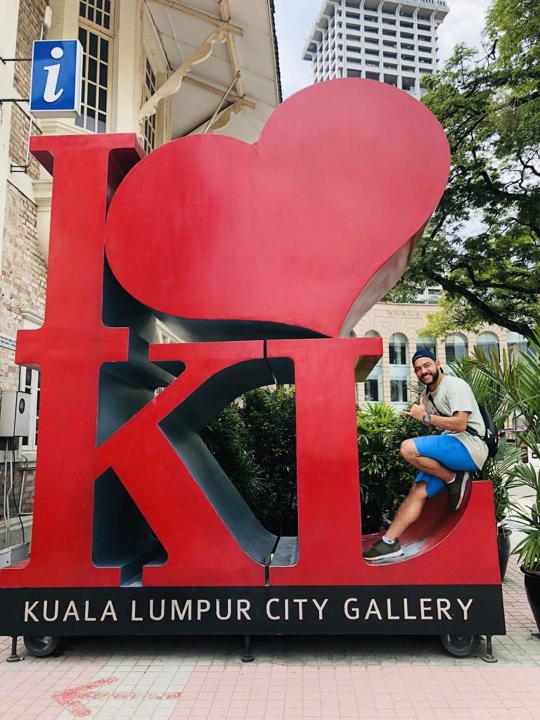 Tiago Lopes sentado no letreiro vermelho I love KL, em Kuala Lumpur