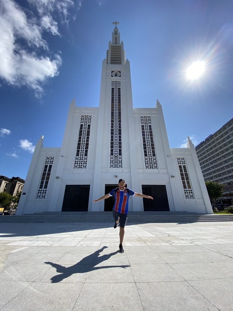 Tiago Lopes saltando em frente à Catedral de Maputo, branca, vestindo a camisa do Bahia