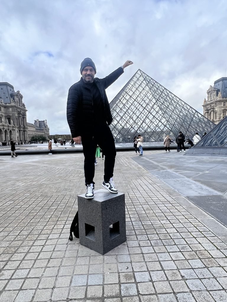 Tiago Lopes em pé sobre um bloco de pedra, com a pirâmide do Louvre ao fundo, em Paris