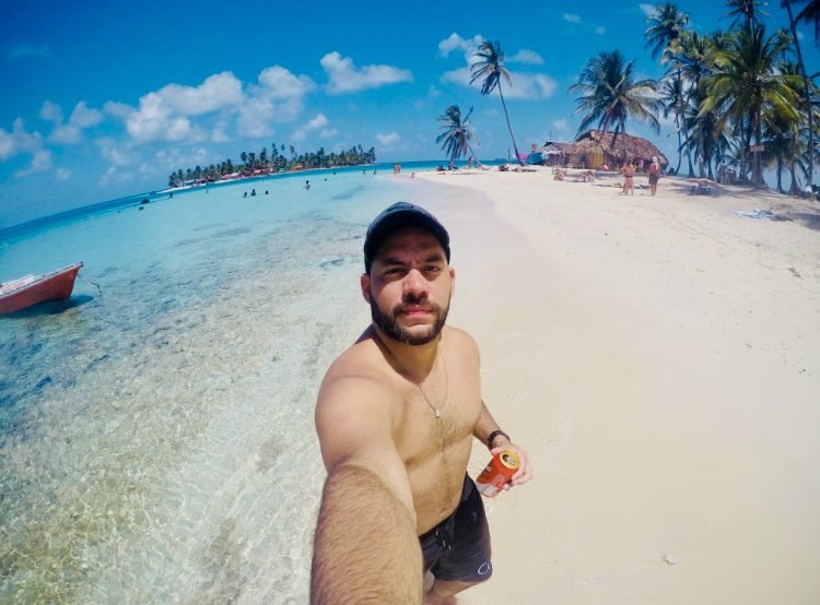 Tiago Lopes em selfie na praia, com cabanas de palha e coqueiros