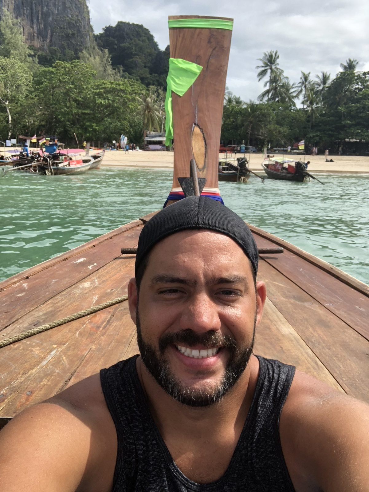 Tiago Lopes sorrindo na proa de um barco long-tail, com praia, coqueiros e falésias ao fundo