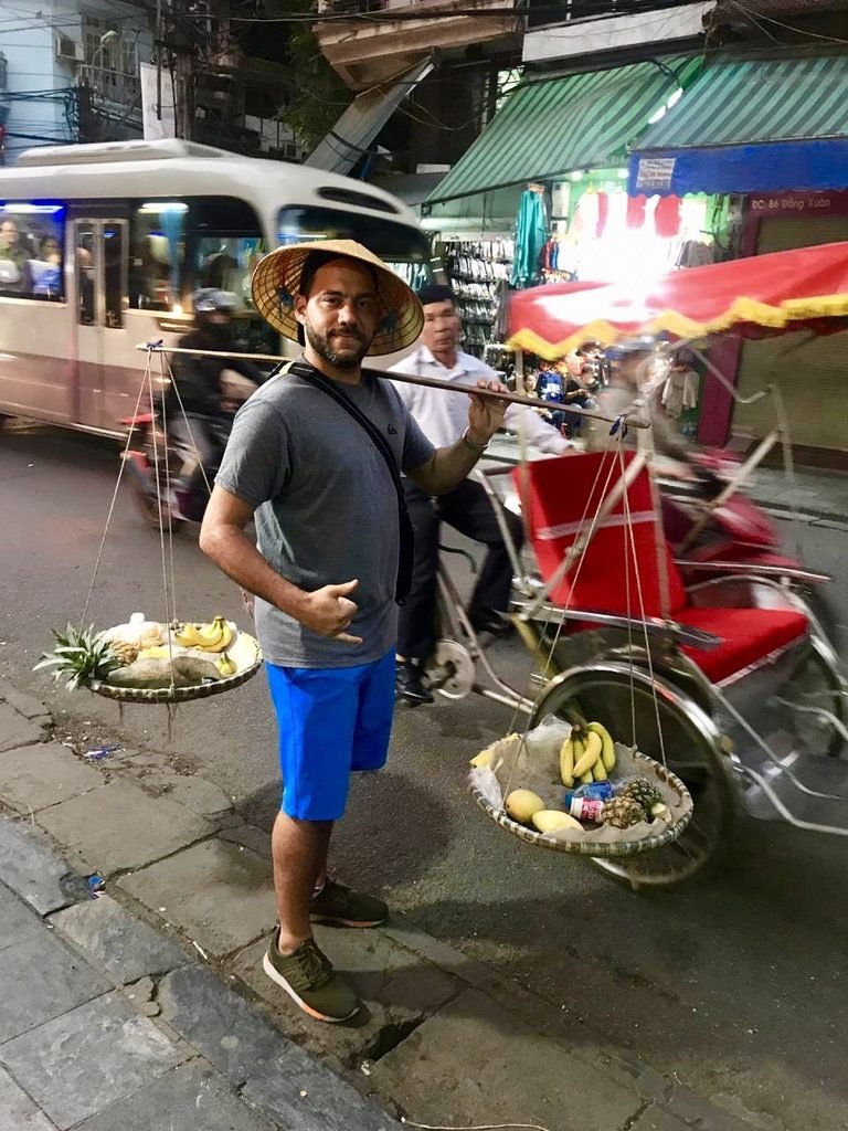 Tiago Lopes com chapéu cônico carregando cestos de frutas numa rua de Hanói, como um vendedor ambulante